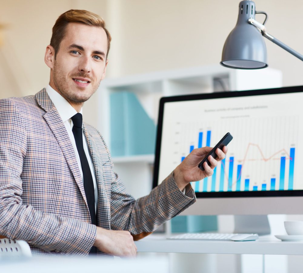 Successful young banker with smartphone sitting by his workplace